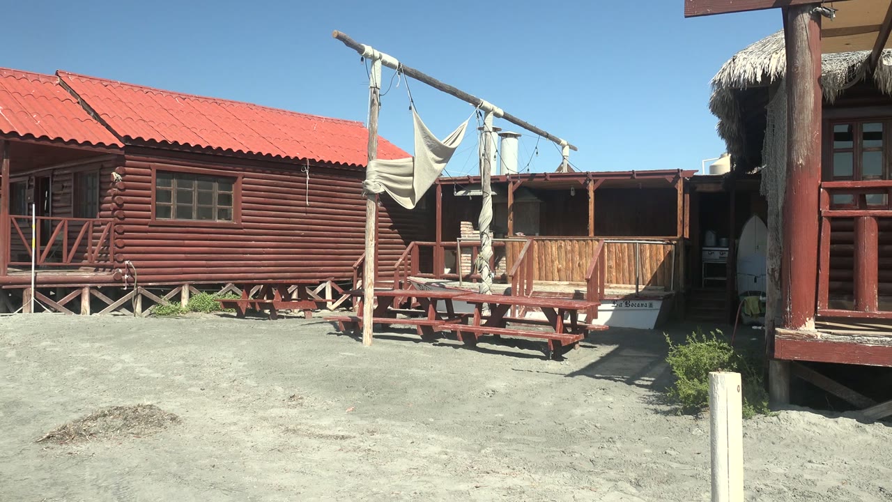 A sunlit La Bocana cabin exterior shows the red metal roof, horizontal wood siding, hammock draped from the frame, picnic table, sandy foreground, and a partly visible figure near the doorway.