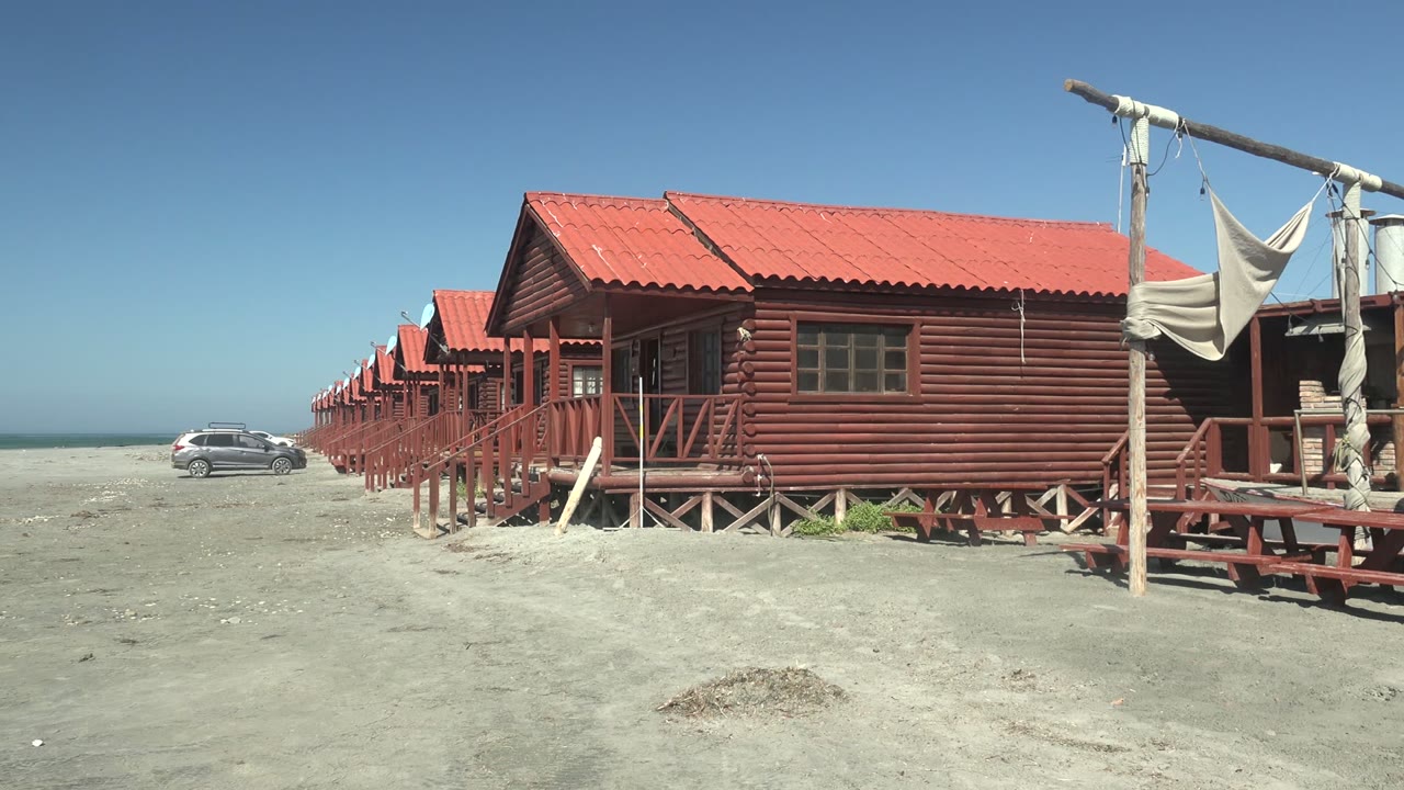 At 0:00 in La Bocana, Baja California Sur, red wooden huts line the sandy beach with a silver car near the ocean and no visible people.