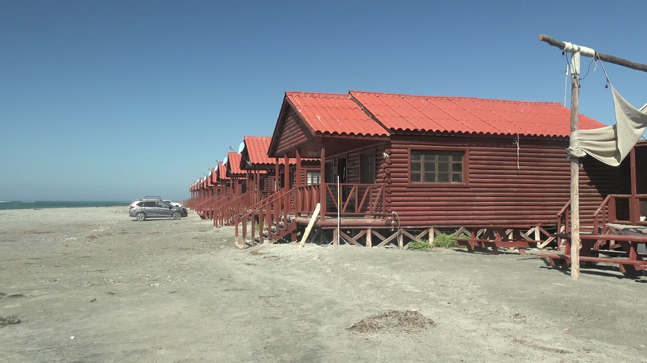 At 0:02, the red cabin fronts and roofline dominate the right side of the La Bocana frame, with pale sand, blue sky, ocean, and no visible people.