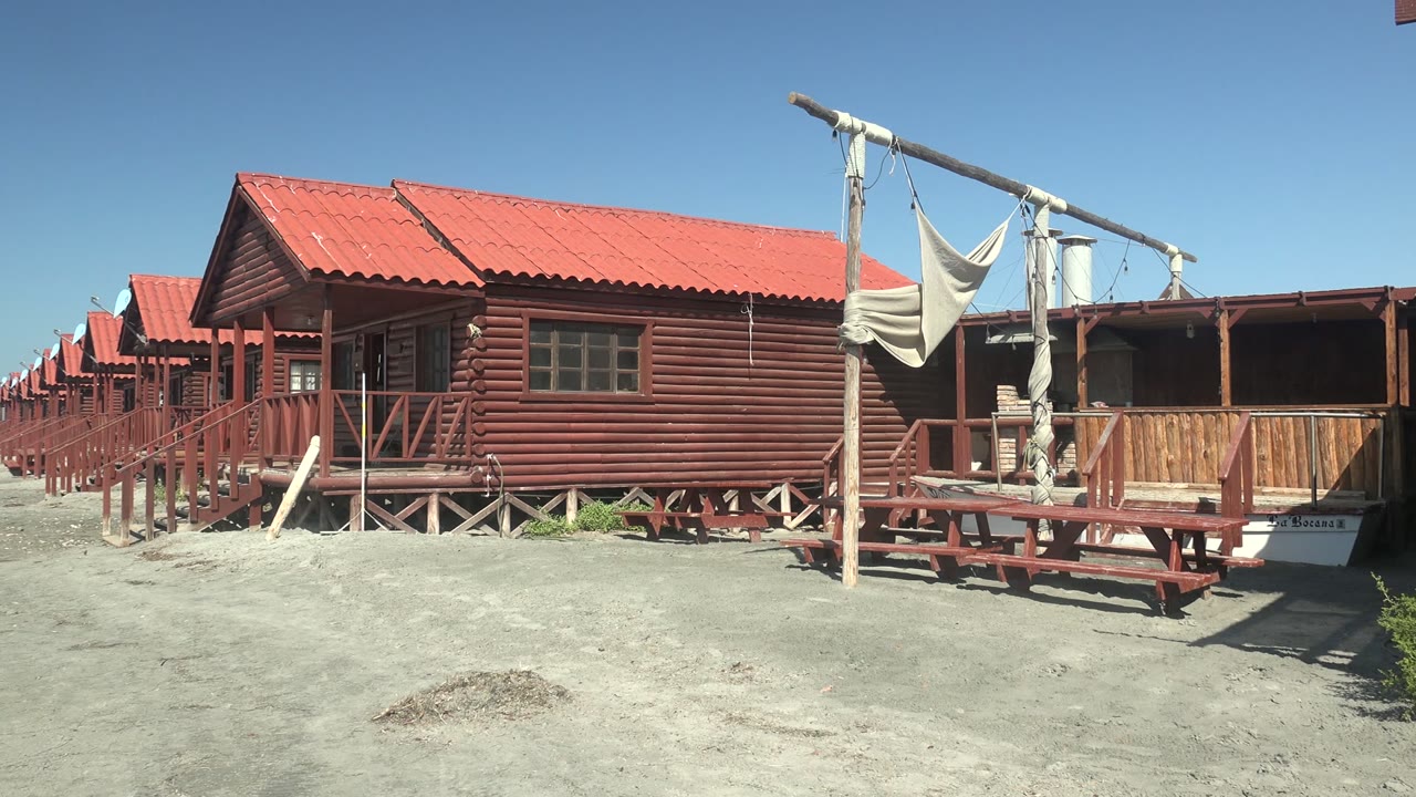 At 0:00 in La Bocana, Baja California Sur, a row of red wooden cabins with red roofs stretches across sandy ground, with porch railings and shade fabric visible and no visible people.