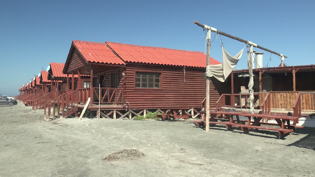 At 0:02 in La Bocana, Baja California Sur, the red cabin row remains in bright sun while a porch frame, hanging shade cloth, picnic table, and water tank occupy the right side with no visible people.