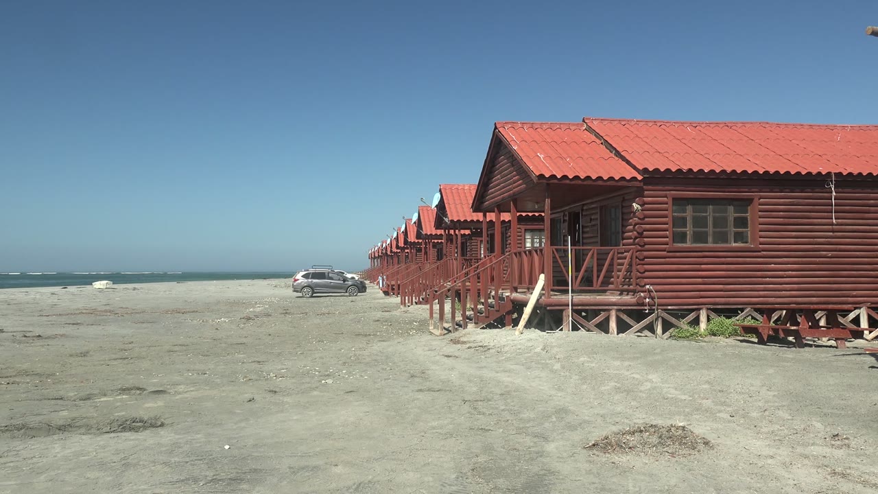 At La Bocana in Baja California Sur, red-roofed wooden cabins line the right side of a sandy beach while a silver car appears near the ocean on the left, with no visible people.