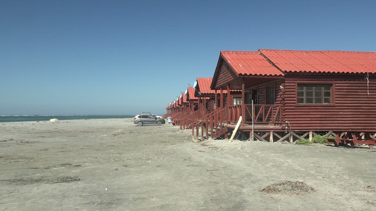 Wooden cabins on a sandy beach with a car closer to the surf.
