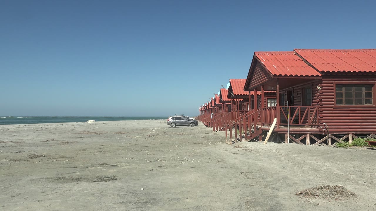 Opening frame in La Bocana, Baja California Sur showing red-roofed wooden cabins on the right, a gray car on the sand near the surf, clear blue sky, and no visible people.