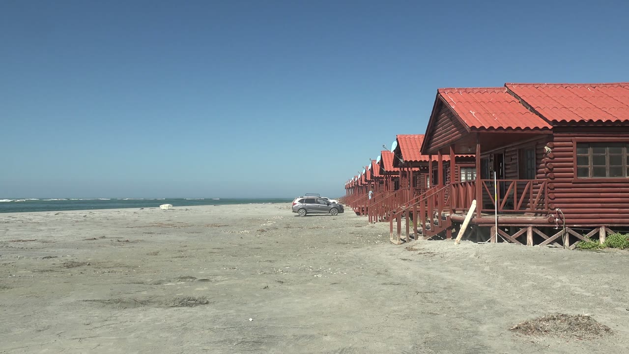Car moving across sand in front of red-roofed beach cabins.