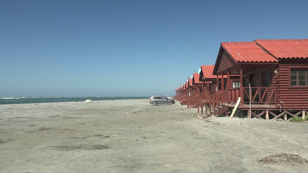 At 0:02 in La Bocana, Baja California Sur, the car continues slowly across the sand near the ocean, with the red-roofed cabins cropped along the right edge and no visible people.