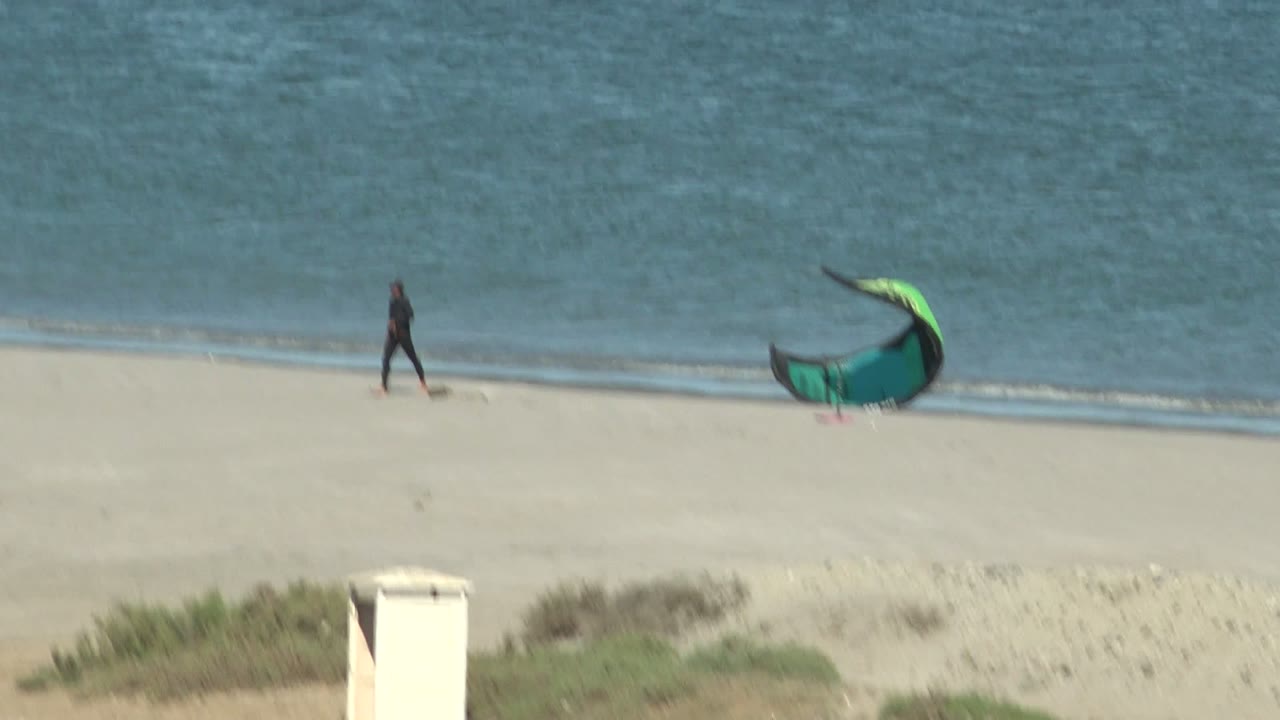 A distant person walks along the sandy beach in San Juanico, Baja California Sur while a green and black kite rests near the shoreline and blue ocean fills the background.