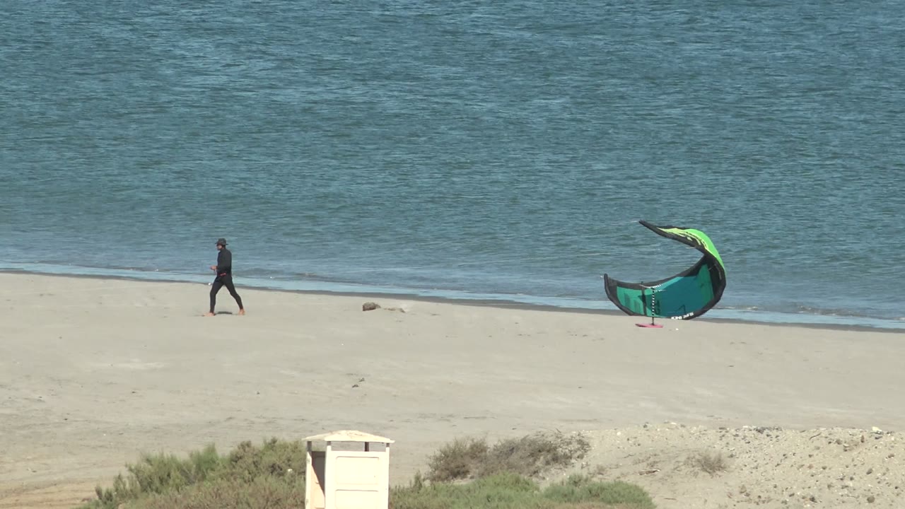 A distant person crosses the pale sand at San Juanico while the green and black kite stands to the right near the waterline, with sparse dune growth below.