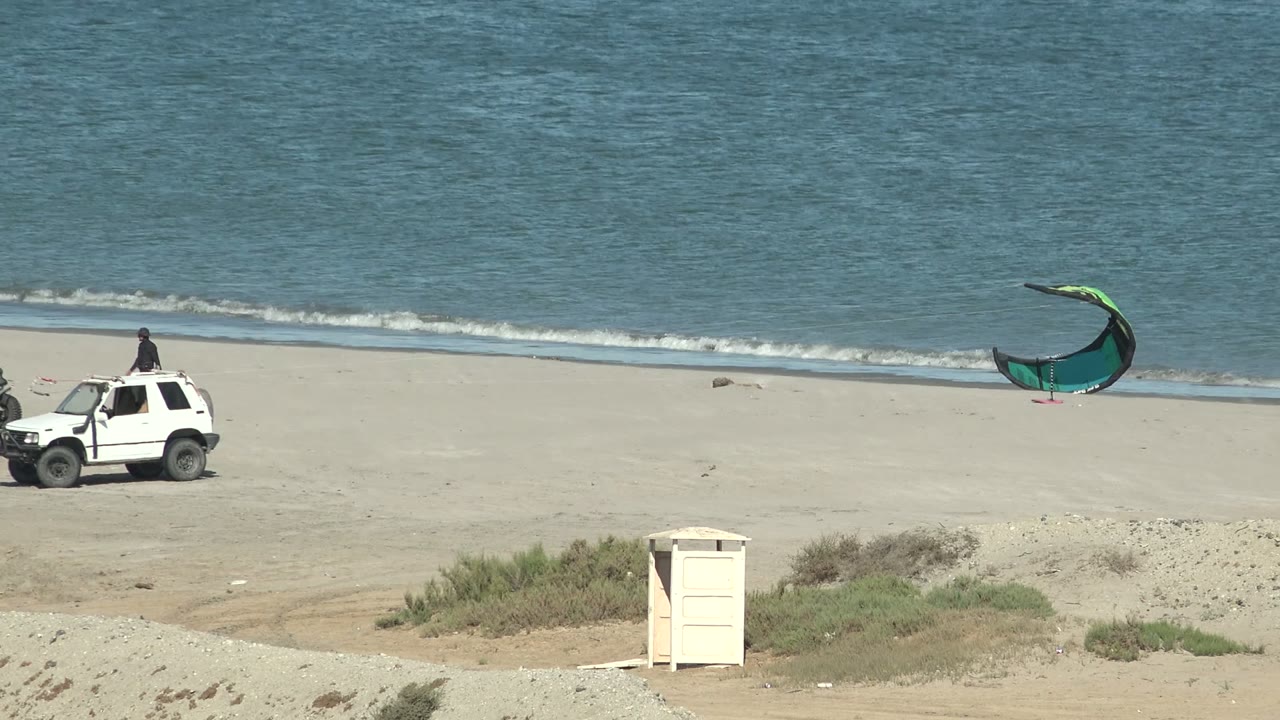 San Juanico, Baja California Sur beach with white vehicles near the shoreline, a green kite by the water, dunes in front, and small people visible near the vehicles.
