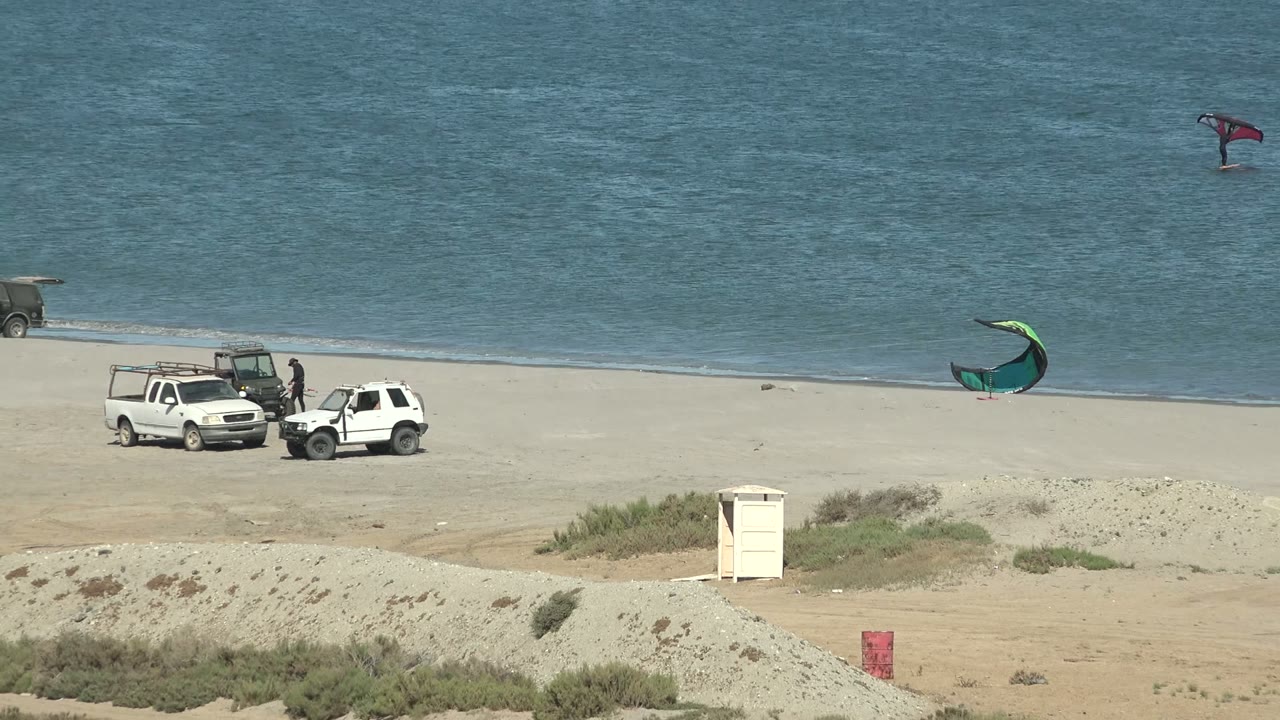 San Juanico beach scene with the vehicles grouped on sand, a green kite near the right shoreline, and a distant red kite surfer high over the blue water.