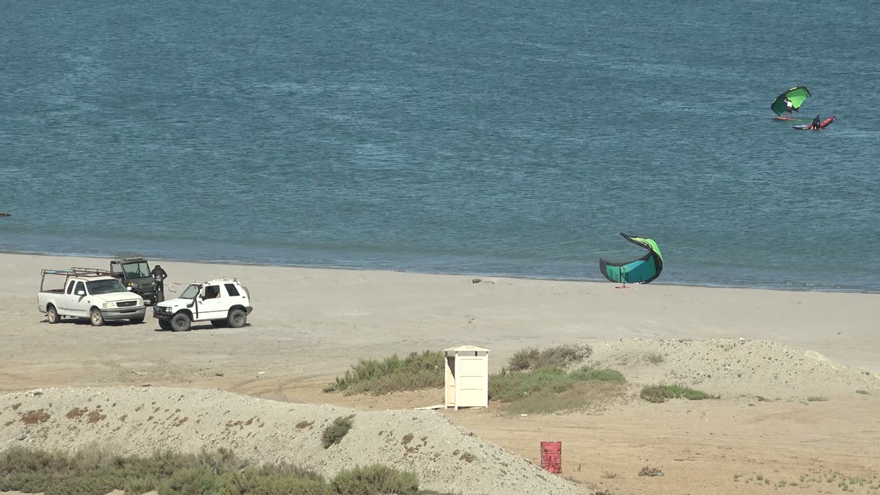 Wide San Juanico beach frame with two white vehicles near the sand, a green kite near shore, and small kite surfers visible on the blue ocean.
