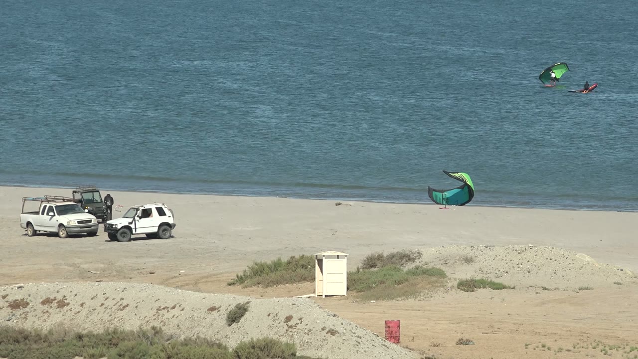 White truck and car on beach with kite surfers behind them.