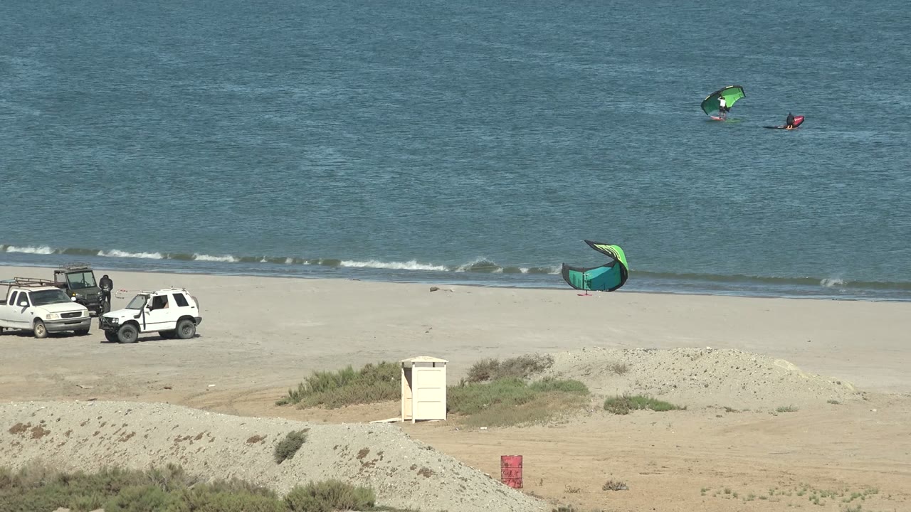 San Juanico beach view with the white vehicles shifting left in the frame, a green kite near the shore, distant kite surfers, and small white wave lines on the water.
