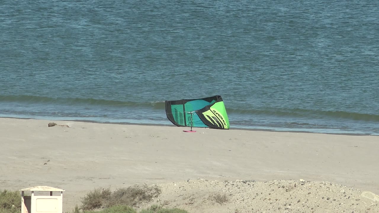 A green and turquoise kite rests on the pale sand at San Juanico, Baja California Sur, with blue ocean and a narrow white surf line behind it, no visible people.