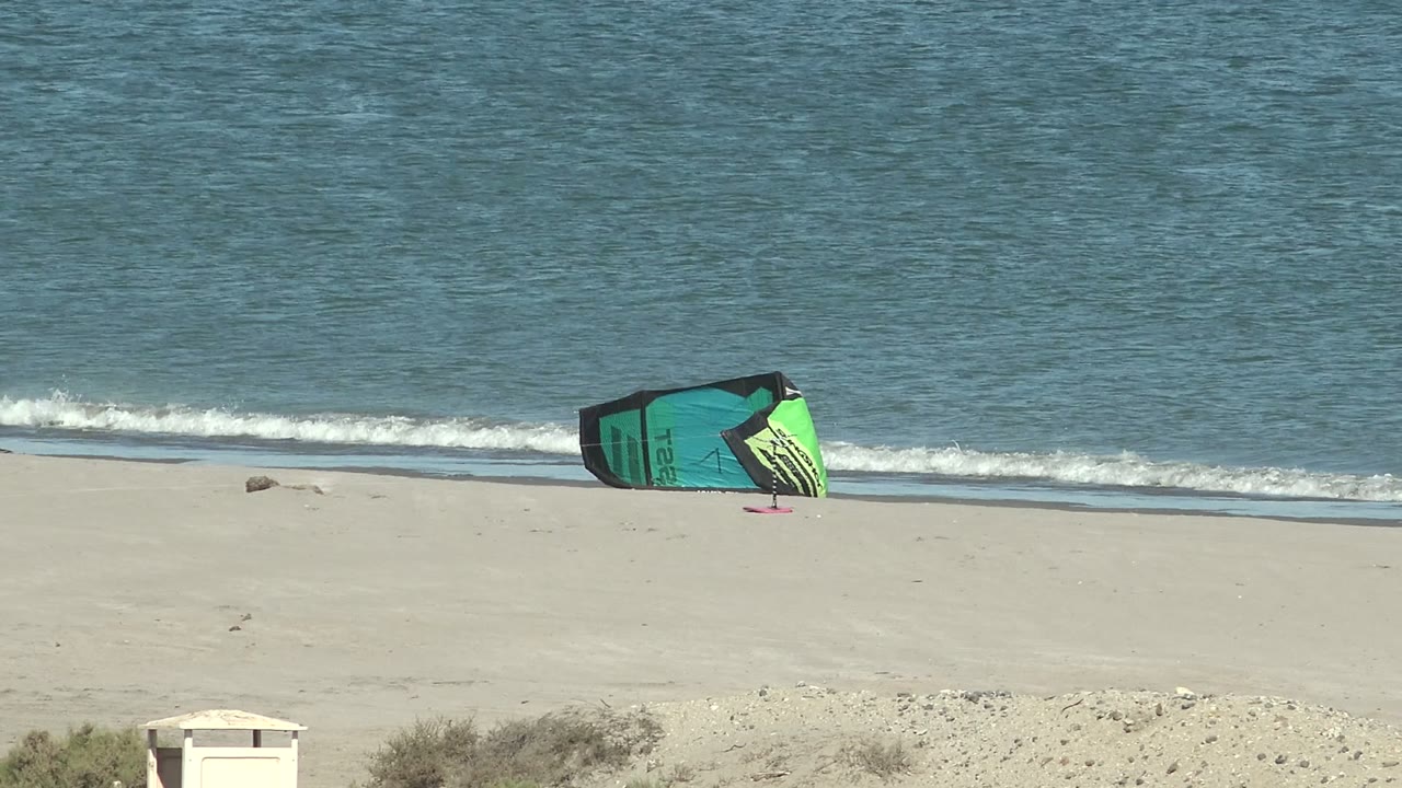 At San Juanico, Baja California Sur, the green and turquoise kite sits on open sand while foamy water spreads along the shoreline behind it, no visible people.