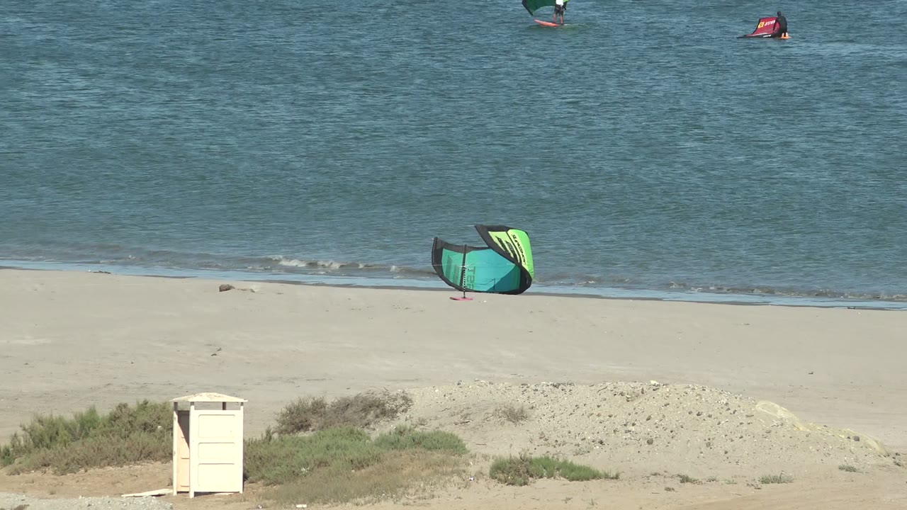 Green and yellow kitesurfing kite sits near the waterline on a sandy beach in San Juanico, Baja California Sur, with blue ocean behind it and no visible people.