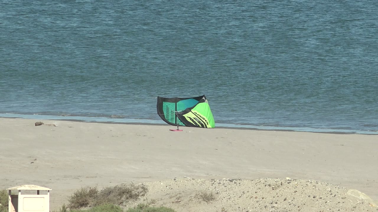 A green and yellow wind kite leans on the sand near the shore in San Juanico, Baja California Sur, with low blue water behind it and no visible people.