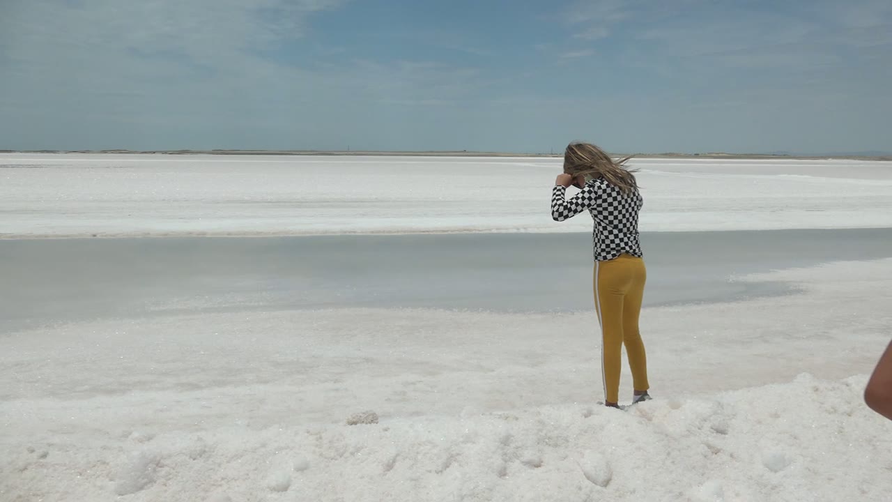 A woman in a checkered shirt and yellow pants faces away while photographing the white flat landscape in Guerrero Negro, Mexico.