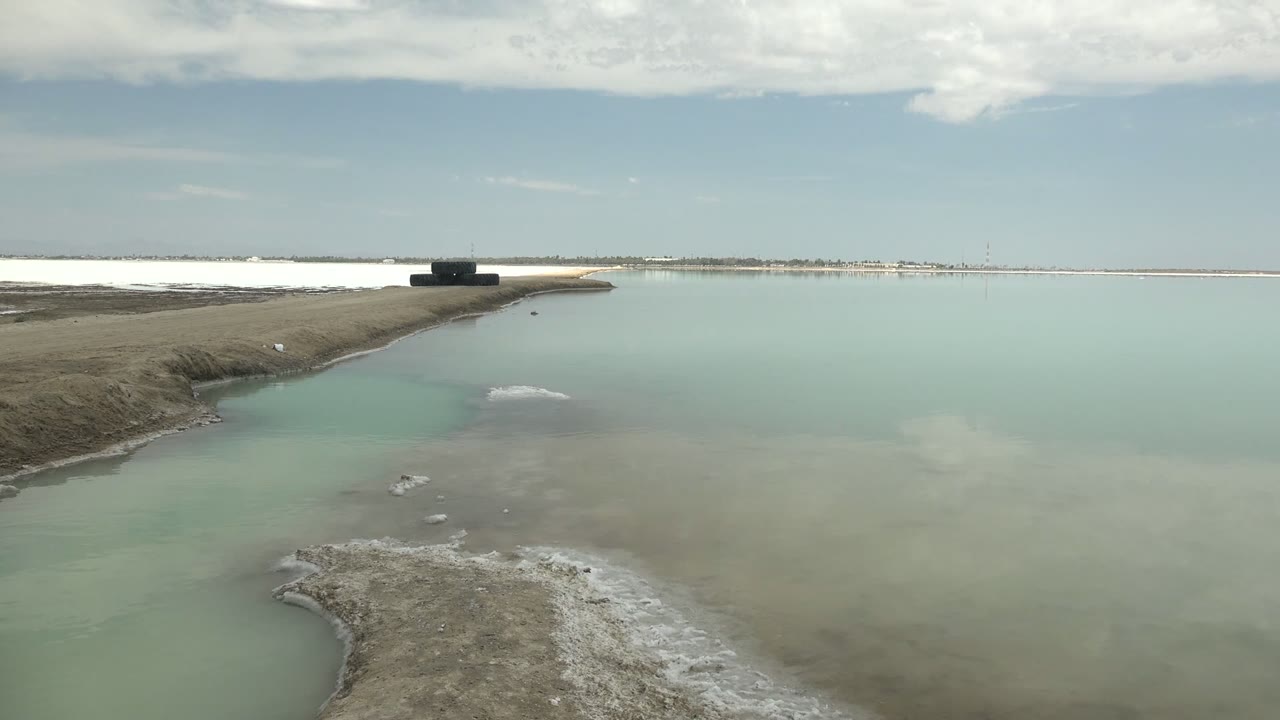 Turquoise water and salt-crusted shoreline in Guerrero Negro, Baja California Sur, with a dark platform on a narrow land strip near the horizon and no visible people.
