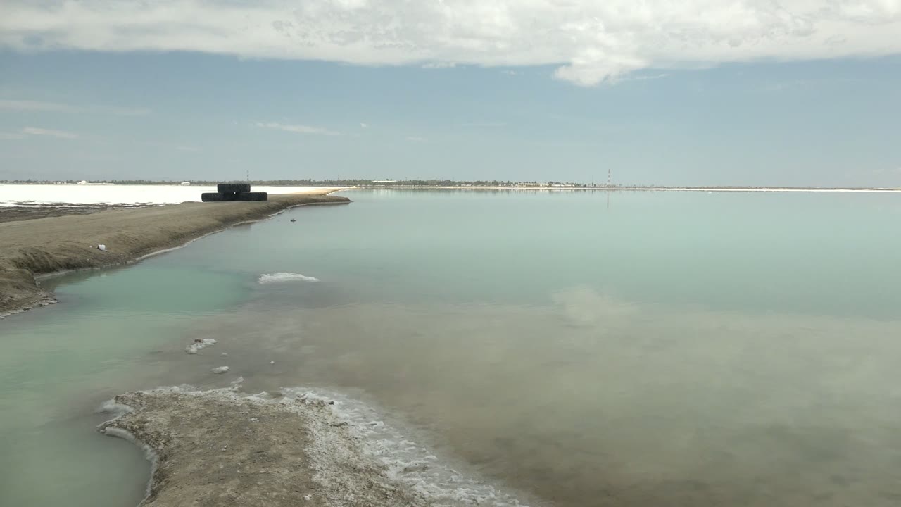 Salt-encrusted ground edges the foreground in Guerrero Negro as pale turquoise water stretches to a thin horizon with a dark platform left of center and no visible people.