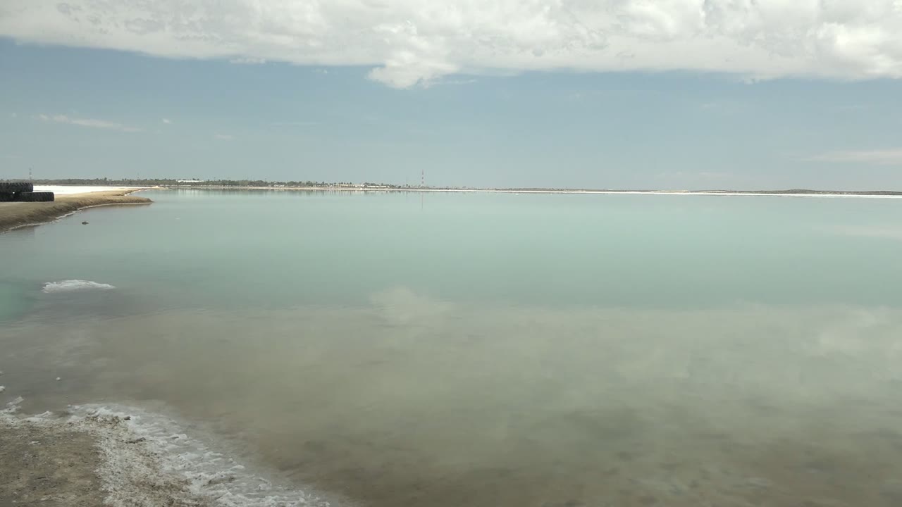 Opening view of a calm Guerrero Negro lagoon with pale blue water, white foam near the foreground shore, a concrete pier at the far left, cloud reflections, and no visible people.