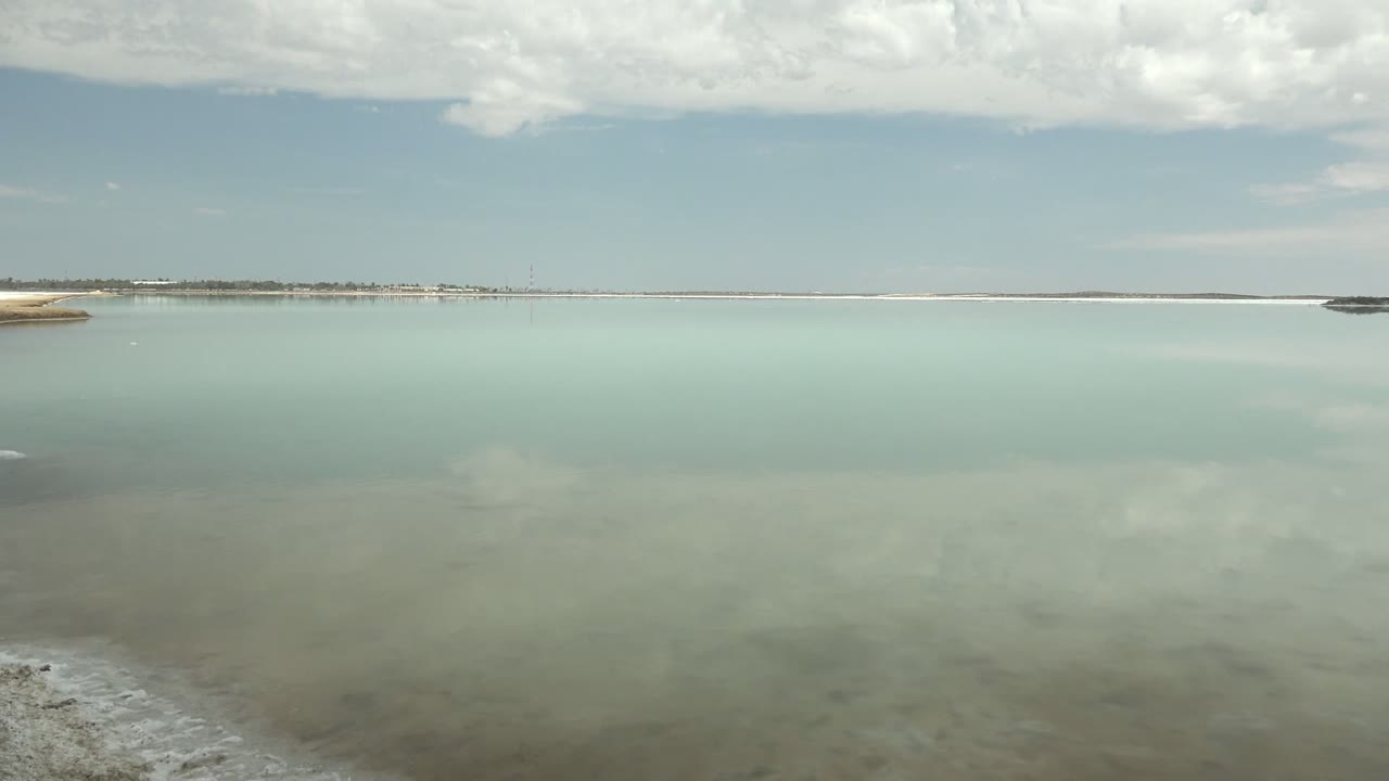 Mid-pan view across the Guerrero Negro lagoon, showing pale green-blue water, a thin distant shoreline, soft cloud reflection across the surface, and no visible people.