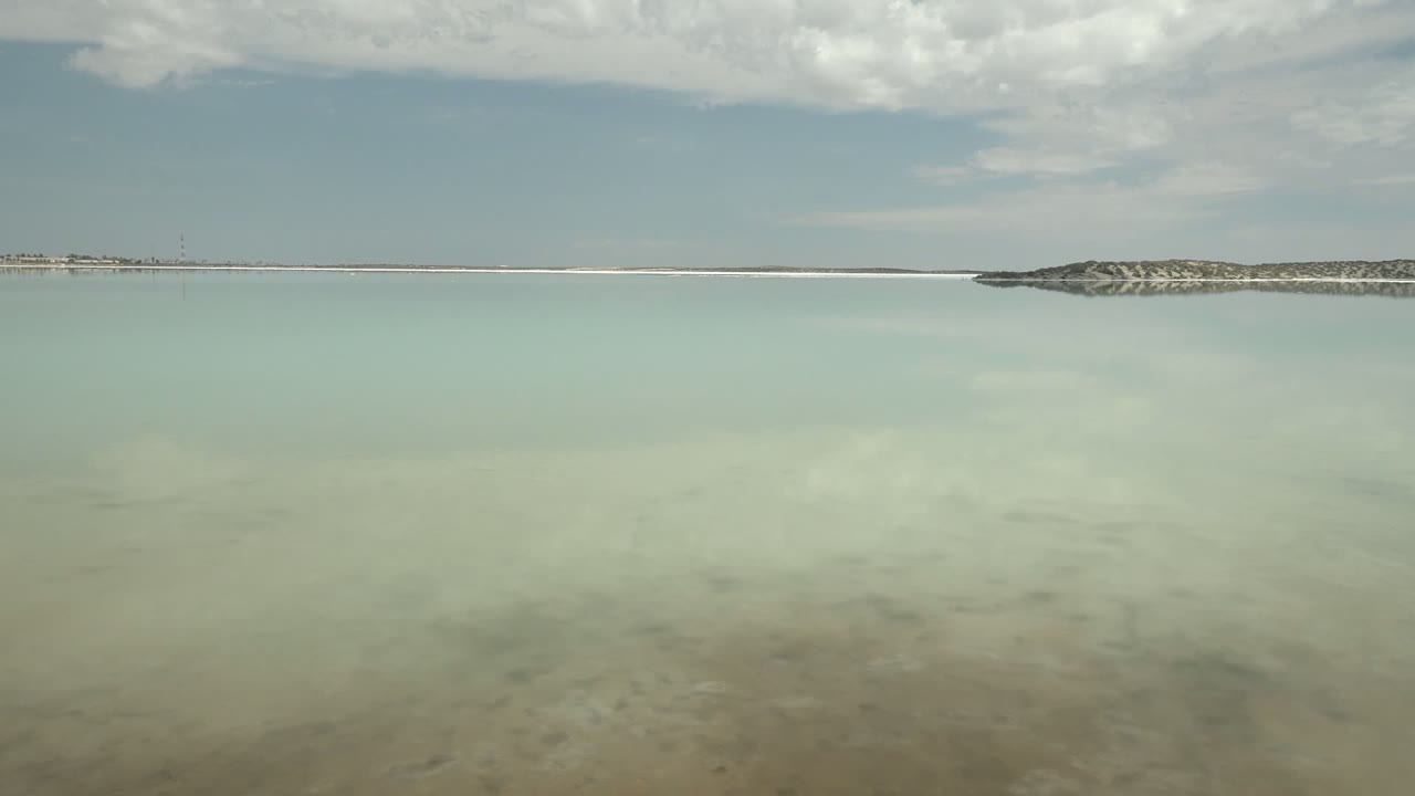 Wide view of a calm salt lake in Guerrero Negro, Baja California Sur, with pale reflective water, a low shoreline near the horizon, cloud cover above, and no visible people.