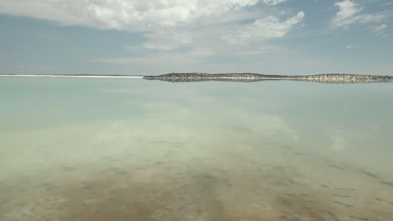 At 0:00 in Guerrero Negro, Baja California Sur, still pale water reflects scattered clouds beneath a low distant shoreline, with no visible people.