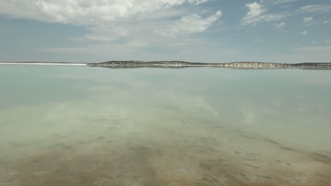 At 0:02 in Guerrero Negro, Baja California Sur, the wide frame shows smooth shallow water, soft cloud reflections, and a low strip of land across the horizon, with no visible people.