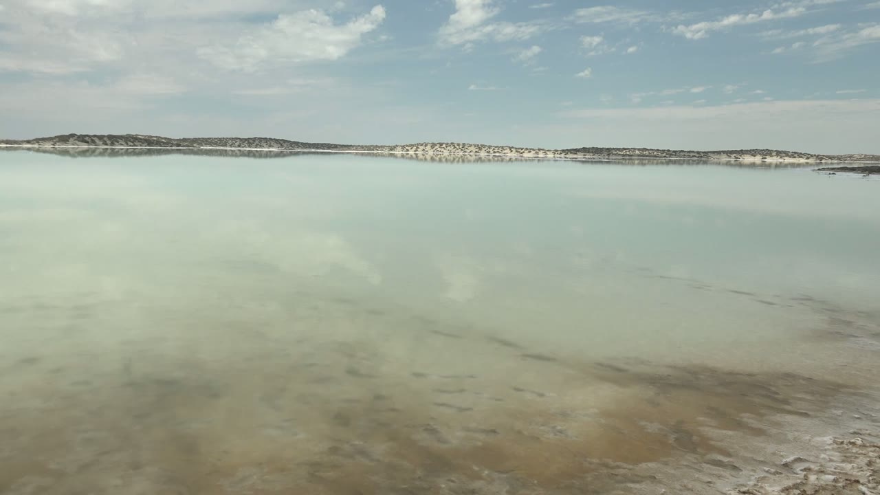 At 0:00, pale shallow salt lake water reflects soft clouds near Guerrero Negro, Baja California Sur, with a white distant shoreline, low hills, and no visible people.