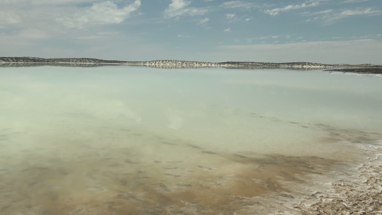 At 0:02, shallow salt water shows darker reflected cloud patches across the foreground while the white shoreline and low hills remain distant, with no visible people.