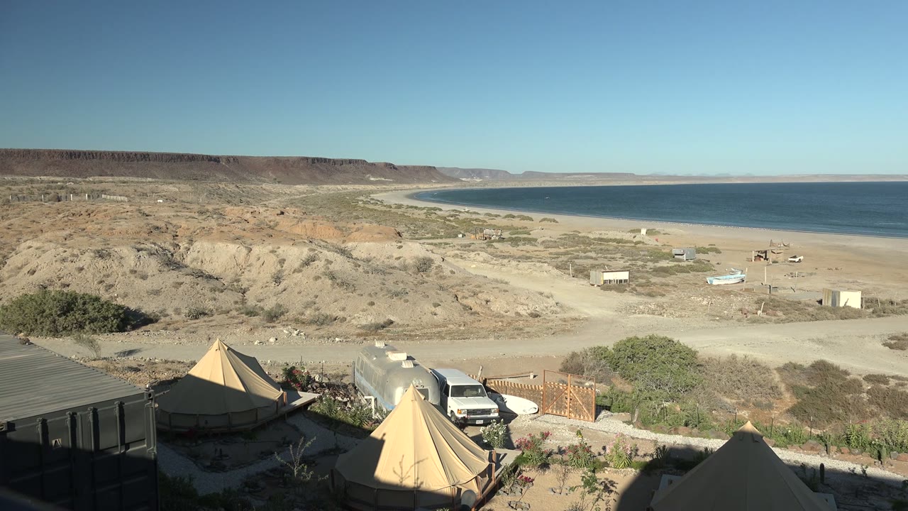 Wide view of a tented camp above the San Juanico, Baja California Sur coastline, with tan tents, a white vehicle, desert scrub, cliffs, beach, blue ocean, clear sky, and no visible people.