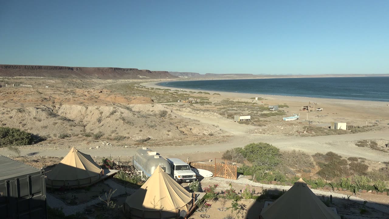 San Juanico desert camp seen from above with tent peaks, a parked white vehicle, a fenced entry, sandy flats, low structures, cliffs, beach, blue water, and no visible people.