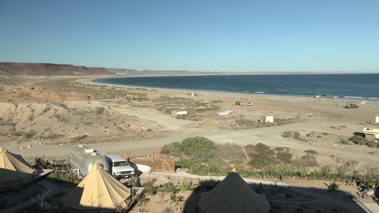 Wide view of a San Juanico desert beach campsite with tent peaks in the foreground, parked vehicles beside shrubs, sandy tracks, the ocean bay beyond, and no visible people.