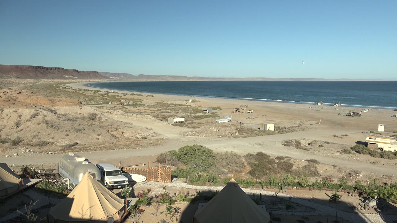 Beach campsite foreground with desert sand, vehicles, and blue ocean.