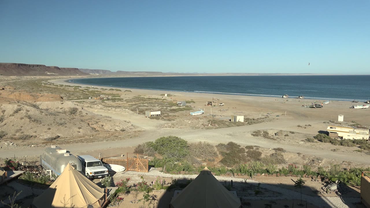 Elevated San Juanico beach campsite frame showing tent tops at the bottom edge, parked vehicles near a fence, sandy roads across the beach, distant boats or small objects by the shore, and no visible people.