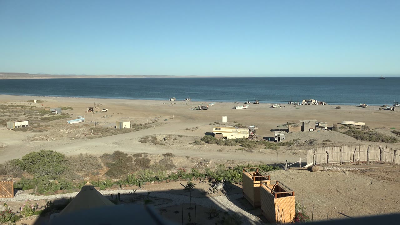 Opening frame of San Juanico, Baja California Sur, showing a wide sandy beach, scattered small structures, blue ocean, clear sky, and no visible people.