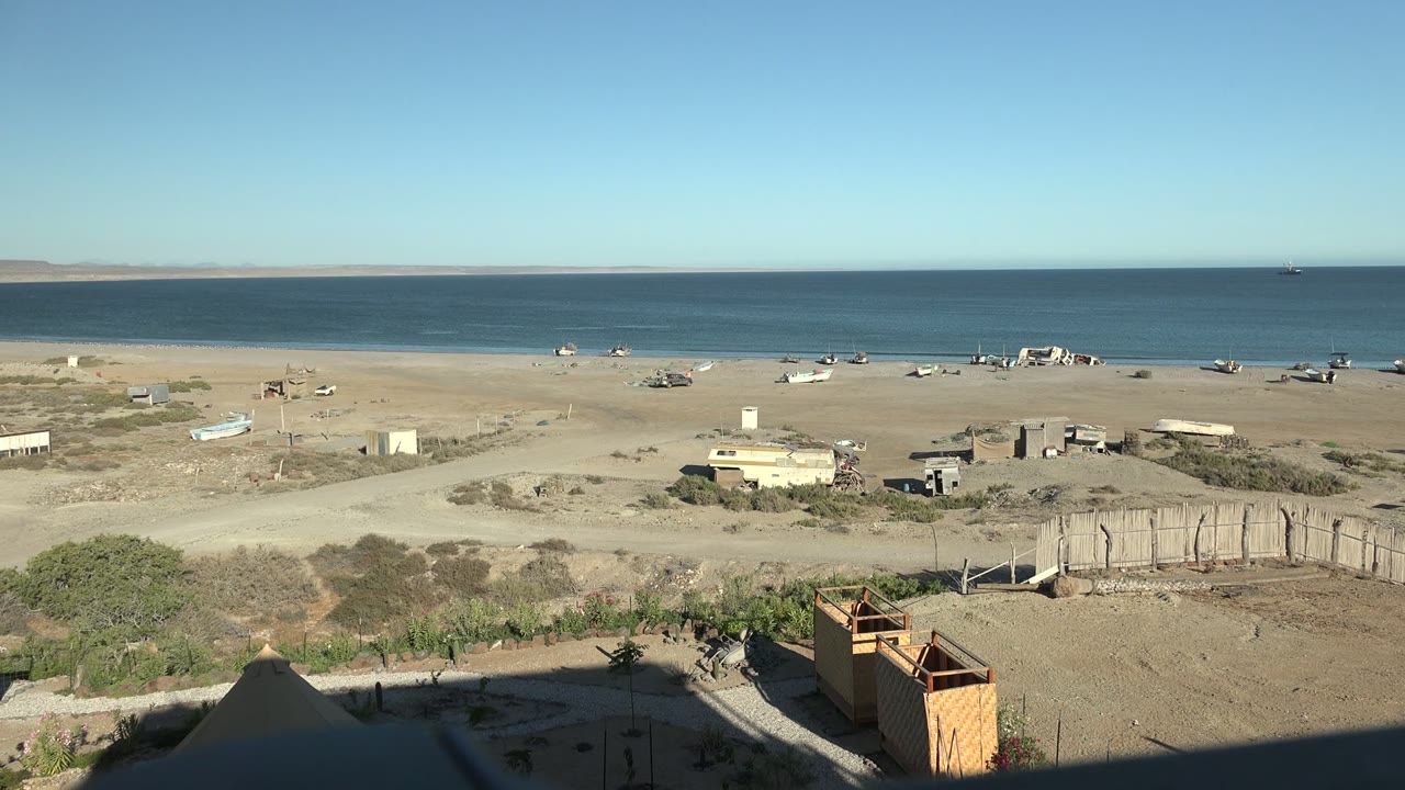 Early frame in San Juanico, Baja California Sur, with the ocean horizon above a dry beach, scattered structures, a wooden foreground element, and no visible people.