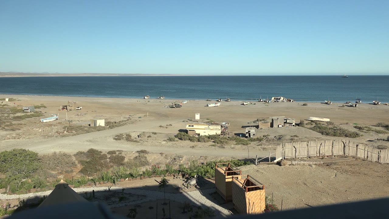 San Juanico beach frame with bright sun on sand, small structures spread across the midground, a dark foreground shadow, blue water, and no visible people.