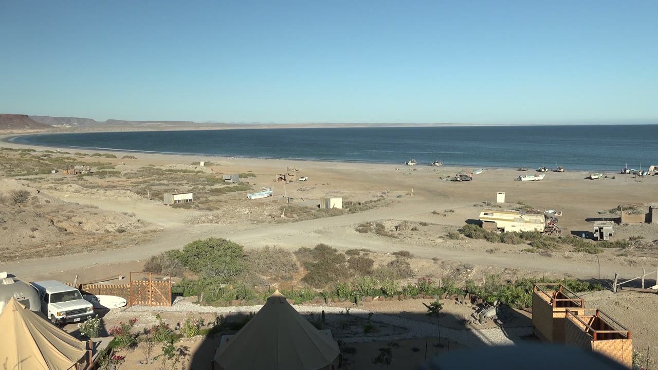 Start of the San Juanico, Baja California Sur clip showing a wide sandy beach, calm ocean horizon, scattered boats, tent tops, wooden fencing, and a white van near the lower left with no visible people.