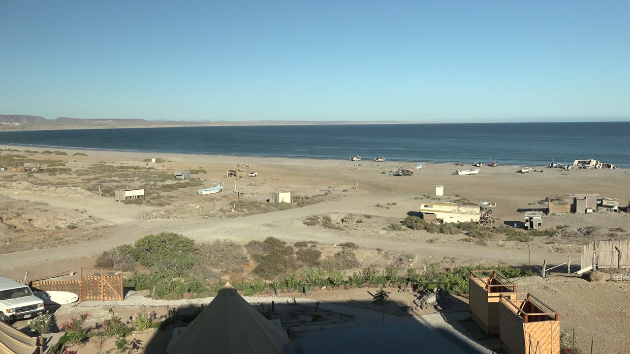 San Juanico shoreline at 0:02 with the white van near the lower left edge, pale sand, scattered boats, low beach structures, blue ocean, distant landforms, and no visible people.