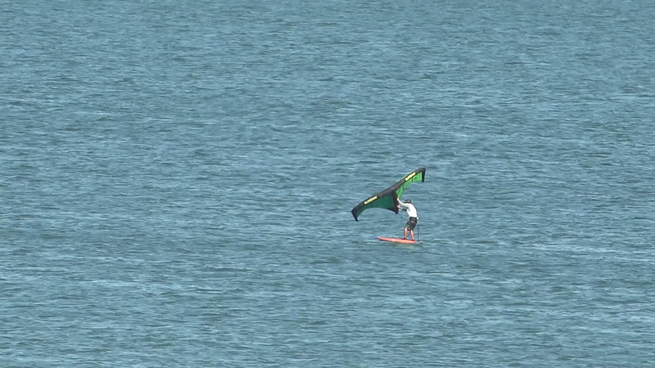 A small distant rider in San Juanico holds the green and black wing high while the red board tracks across lightly rippled blue ocean water.