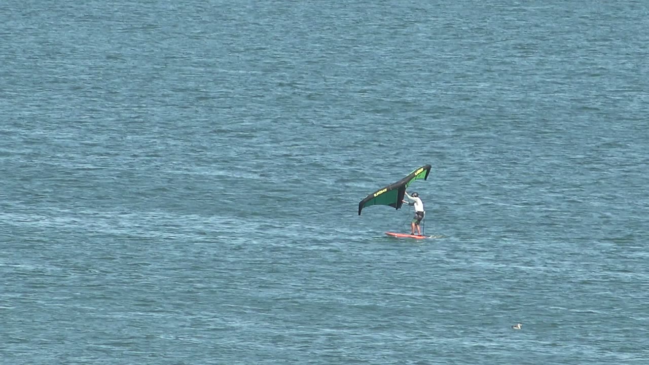 At 0:00, a distant kite surfer in San Juanico, Baja California Sur rides a red board while holding a green and black wing over lightly rippled blue water.