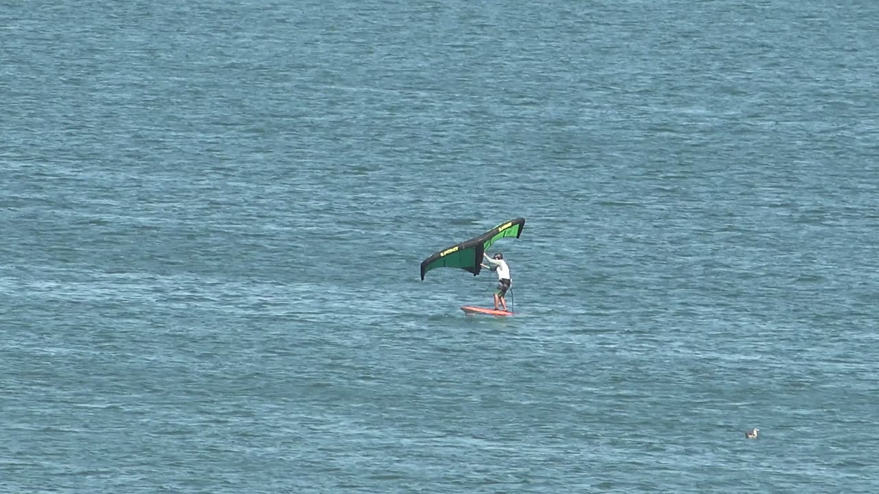 At 0:02, the rider appears farther right in the frame, still balancing on a red board beneath the green and black wing over the blue ocean surface in San Juanico.