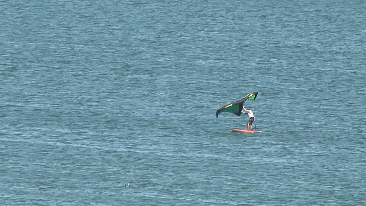 A distant kiteboarder rides a red board across blue ocean water in San Juanico, Baja California Sur, holding a dark kite with green accents near the start of the clip.