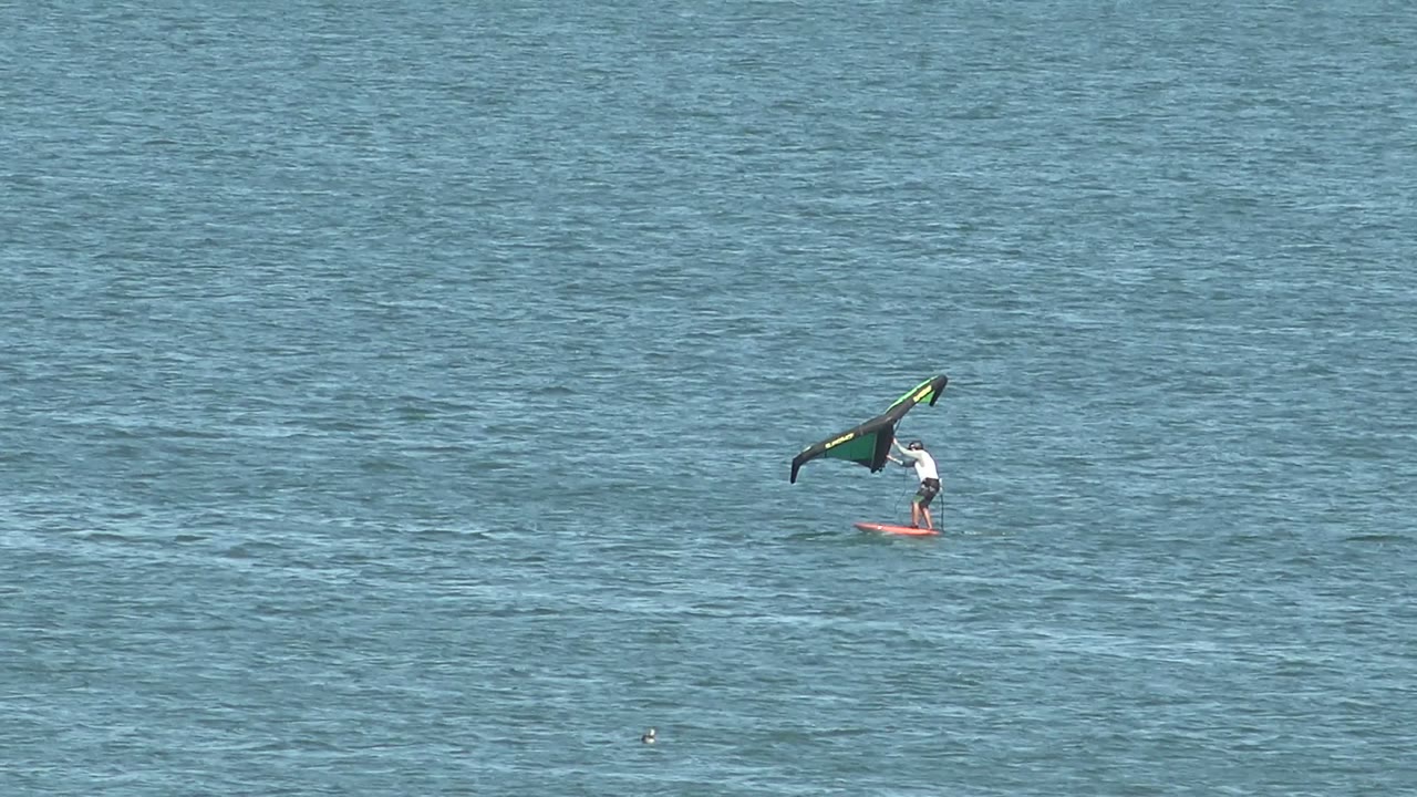 A distant kiteboarder appears farther right over the blue sea in San Juanico, Baja California Sur, with the kite still raised and the board tracking along the water surface.