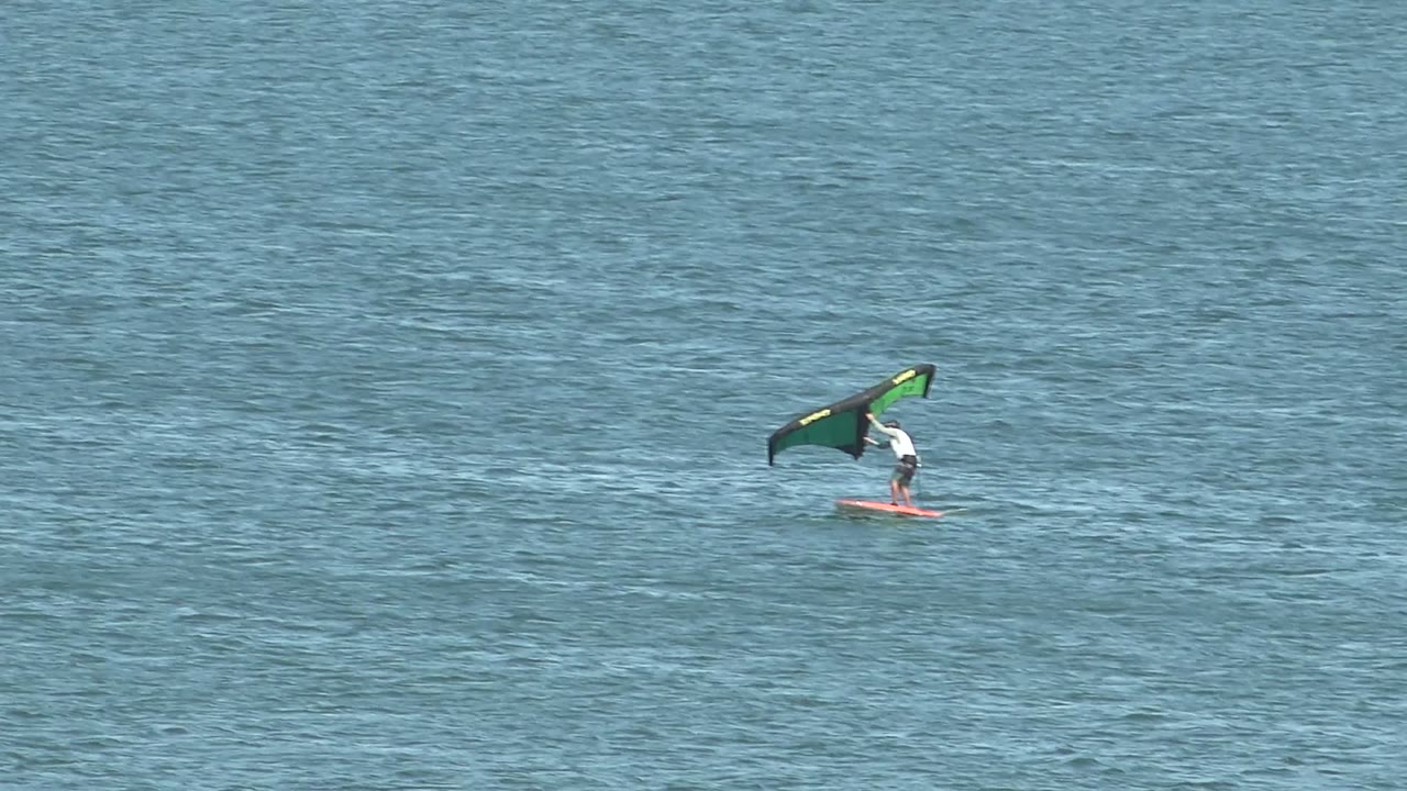 A distant kite surfer in San Juanico, Baja California Sur rides a red board across blue water while holding a green and black kite near the left side of frame.