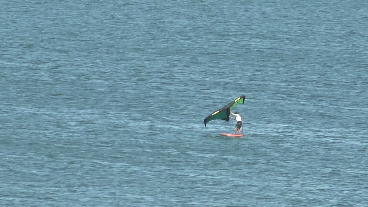 A distant kite surfer in San Juanico, Baja California Sur rides near the right side of the frame, with a green and black kite angled over rippled blue water.