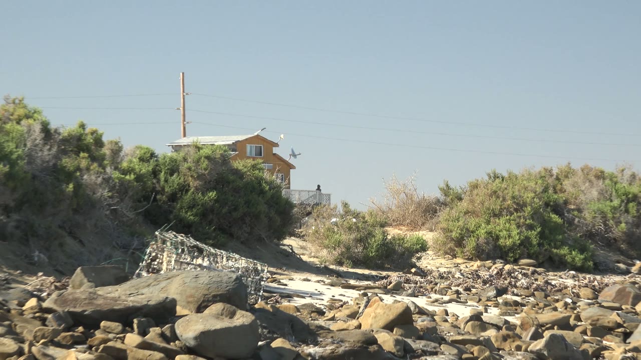 Opening frame of a La Bocana rocky beach with large foreground stones, pale sand, scrub vegetation, a distant house and utility pole, and no visible people.