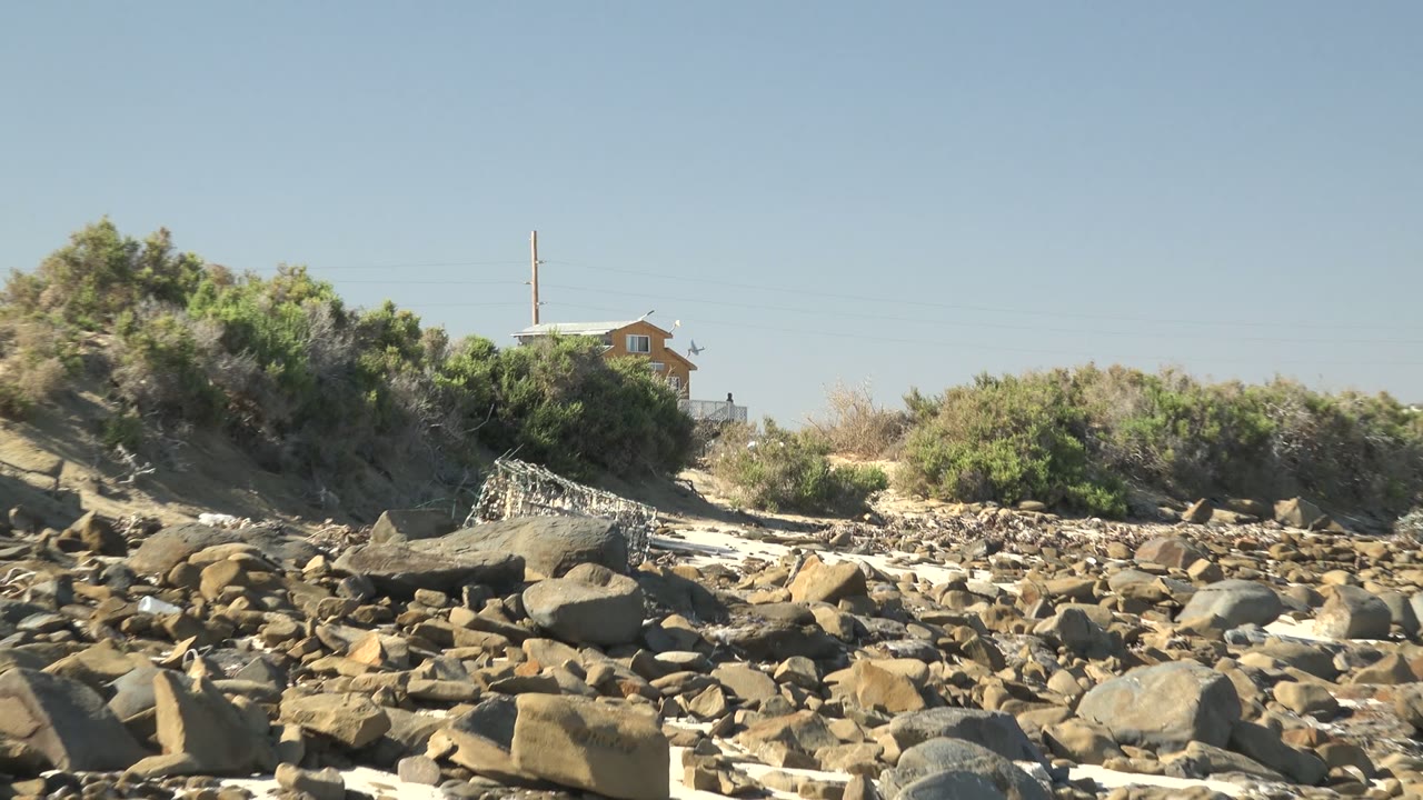 Rocky sandy shore with low green vegetation and a small distant house near the top of the frame.